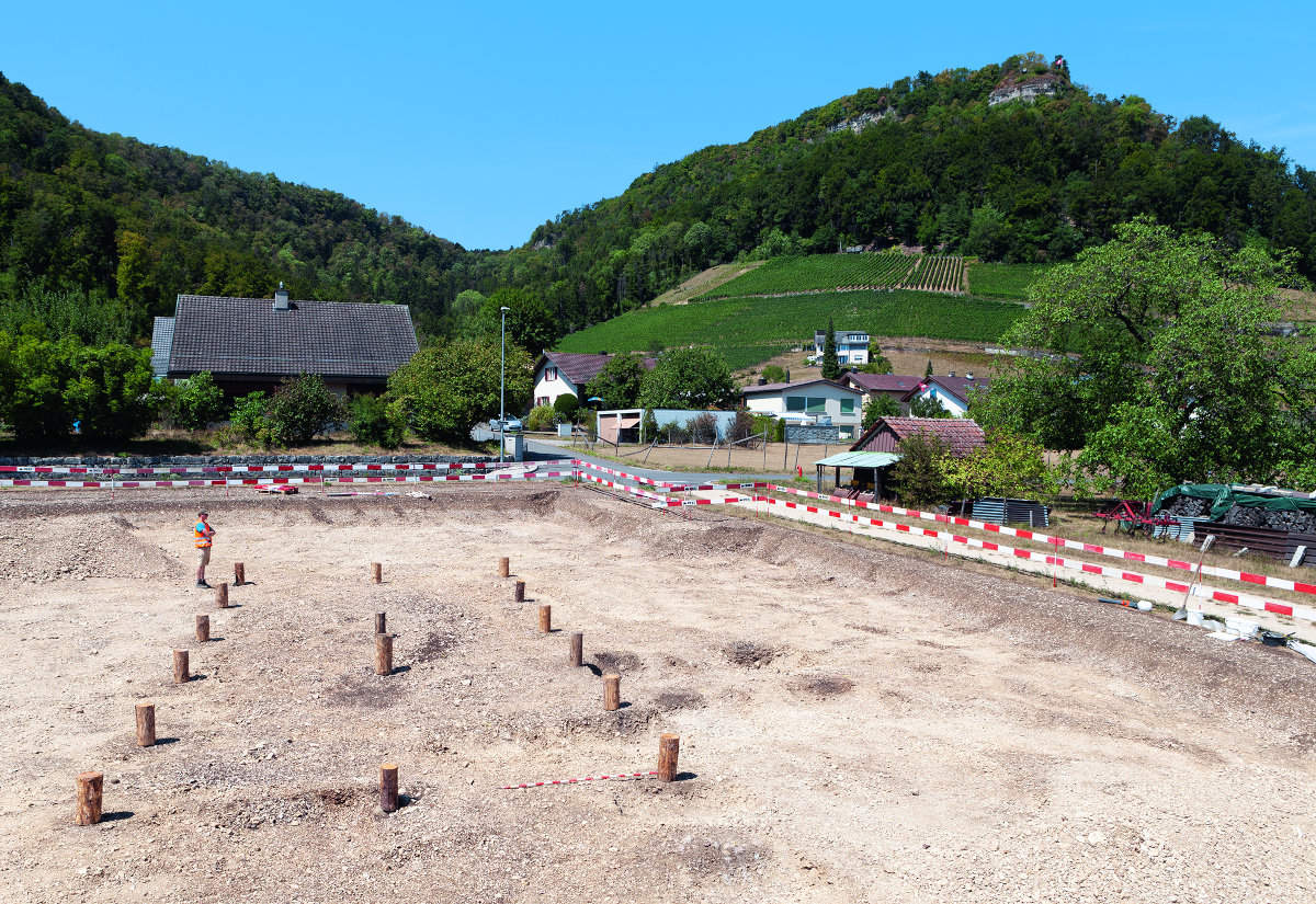 Villigen im Kanton Aargau. Siedlung der Mittelbronzezeit mit einem Haus von 12 m Länge und 6,5 m Breite. Für das Foto wurden Holzklötze in die Pfostengruben gestellt. Im Hintergrund der fel - sige Geissberg. Hier lag eine defensive Siedlung der Spätbronzezeit.