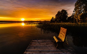 Panoramaaufnahme von einem Sonnenuntergang vor einem See mit einer Bank