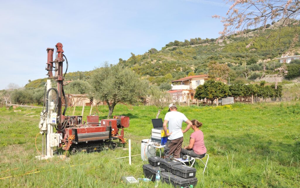 Zu sehen sind zwei Forscher, die auf einem Feld in der Nähe des archaischen Tempels bei Kleidi stehen bzw. sitzen. Vor ihnen befindet sich ein Laptop sowie eine Maschine für den Einsatz des Direct-Push-Systems.