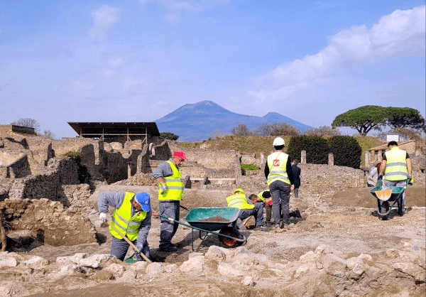 Zu sehen sind mehrere Personen, die zwischen bereits freilegten Mauerzügen in der Regio 9 stehen und weiter an dem Abtransport der Erde arbeiten. Im Hintergrund ist der Vesuv sowie bereits weitere freigelegte Mauern in Pompeji zu sehen.